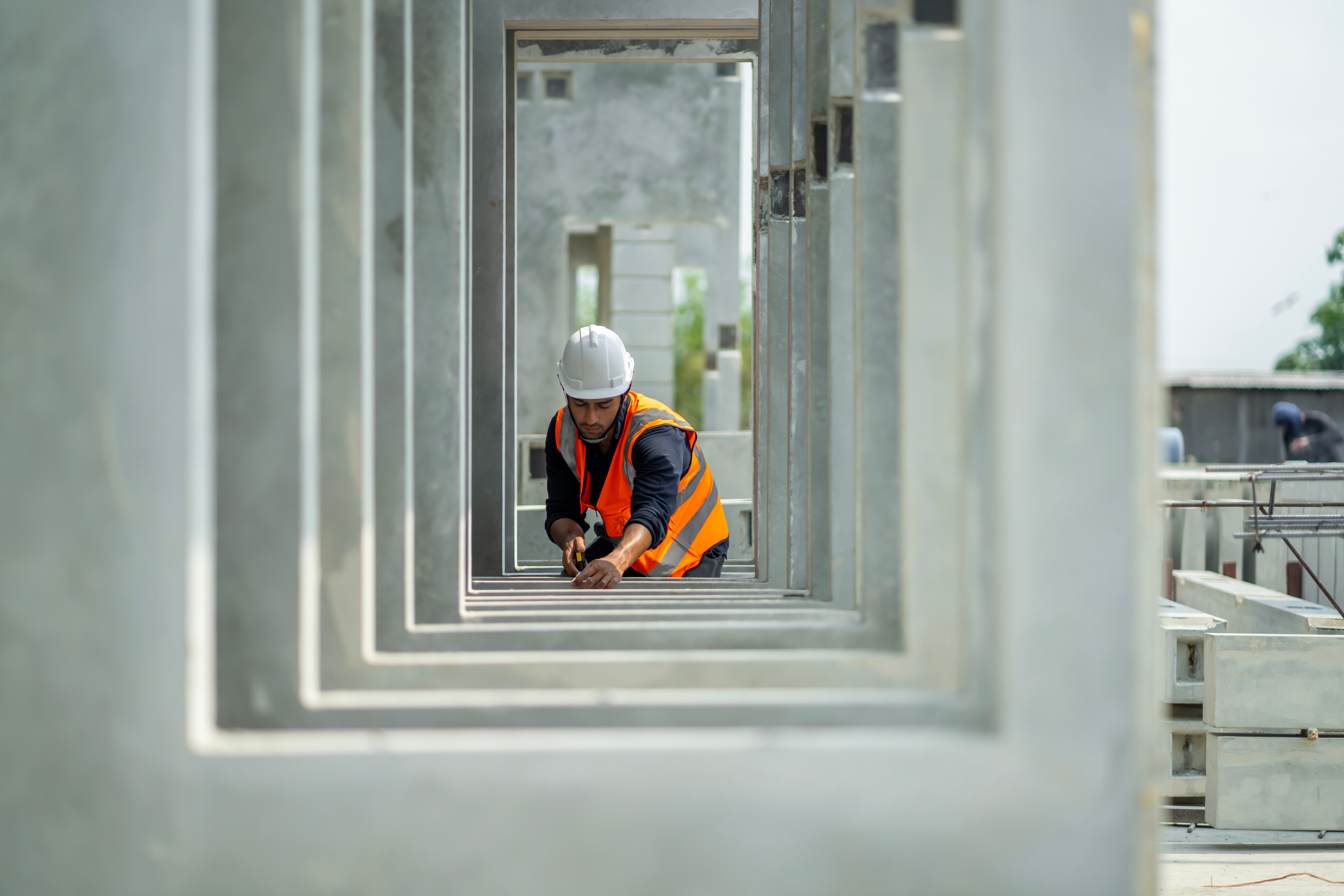 A construction worker wearing a white hard hat and orange safety vest is measuring within a concrete structure.