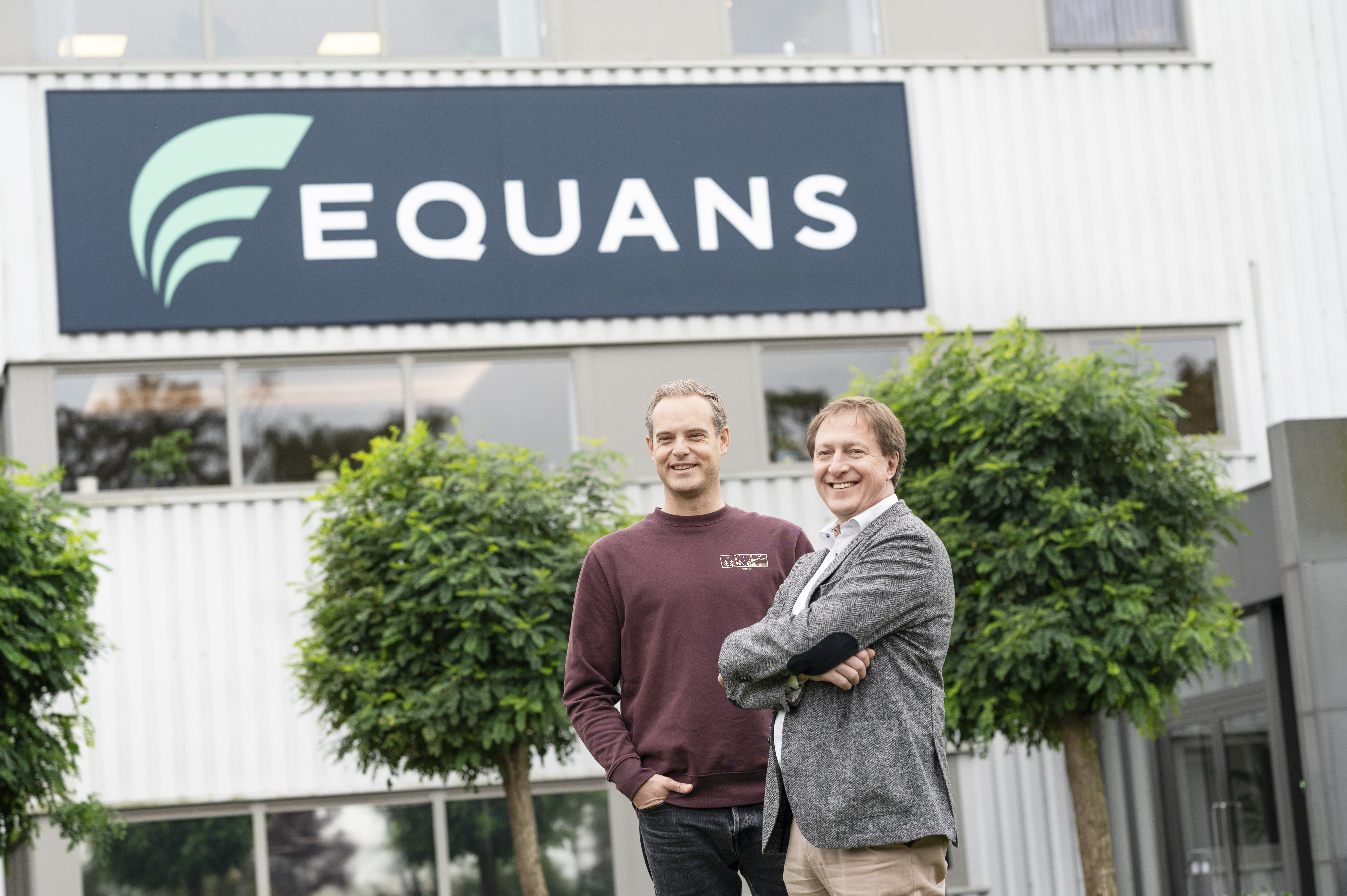Two men standing outside Equans building, with Equans logo prominently displayed above them