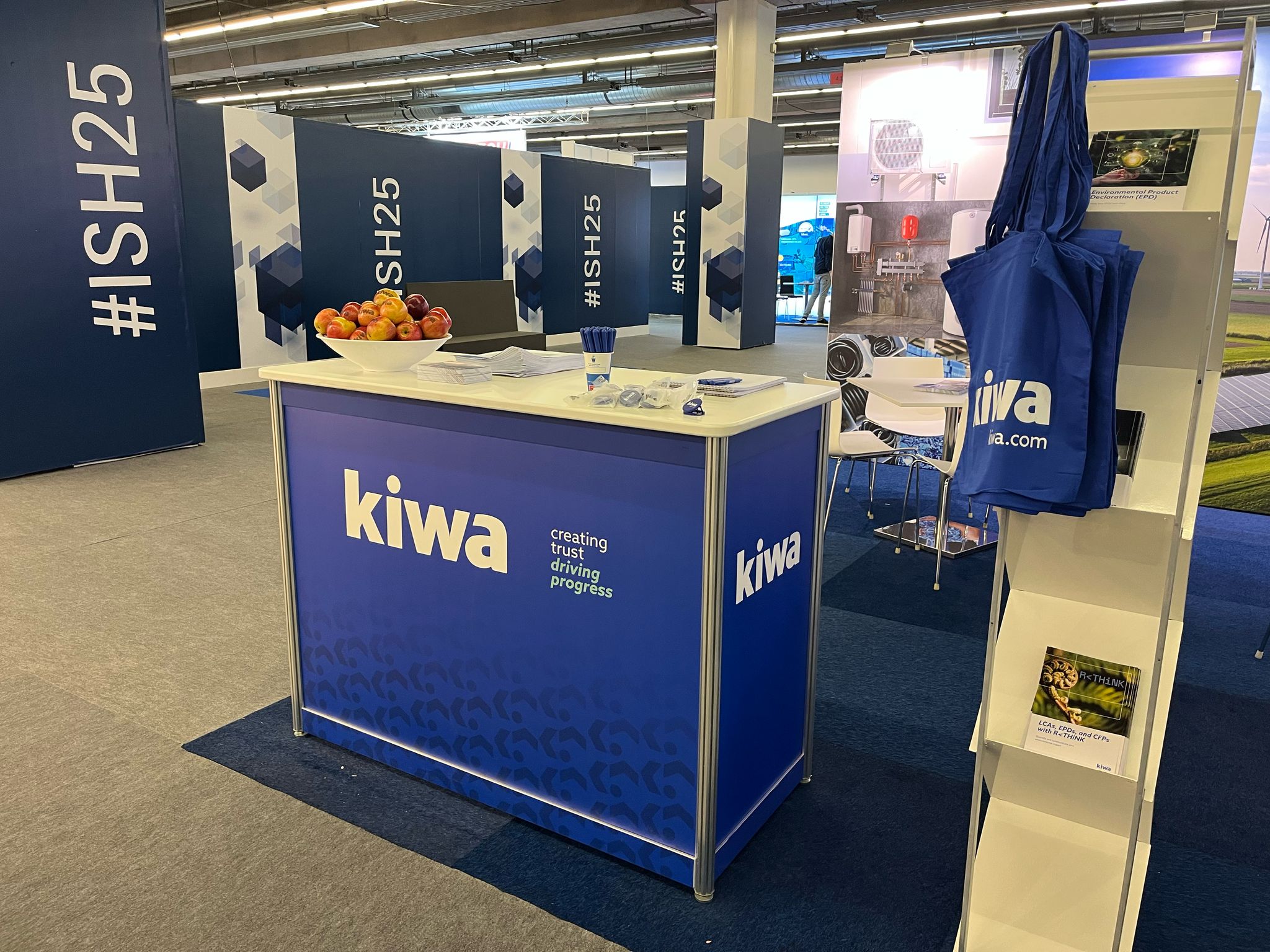 Exhibition booth for Kiwa at ISH25, featuring branded materials, a bowl of fruit, and promotional bags