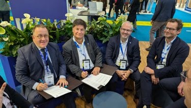 Four men in business suits seated at a table, smiling and holding documents, surrounded by greenery and a busy conference setting