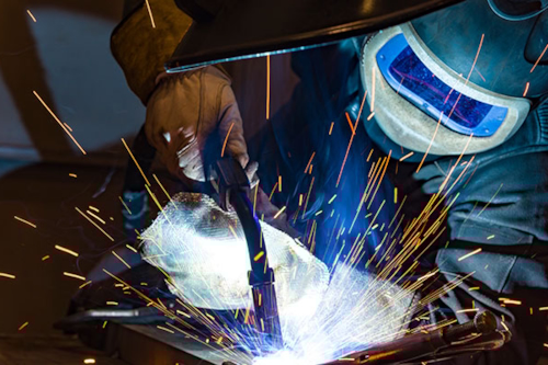 Svetsare lutar lite åt sidan medan han svetsar. Svetslågor sprutar upp.