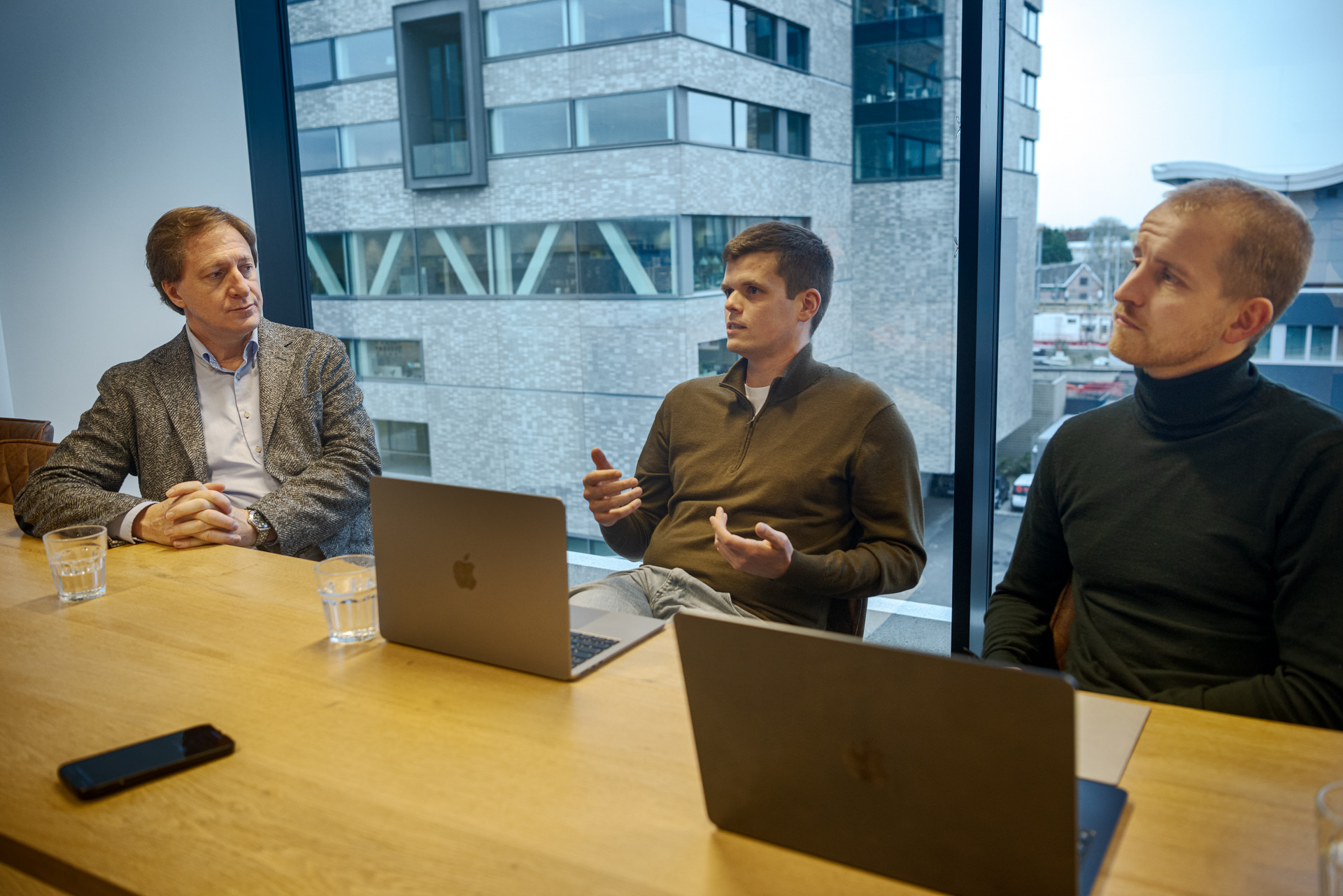 Three individuals seated at a wooden table in a modern office setting, with laptops, glasses of water, and a smartphone on the table; large windows reveal an urban cityscape outside.