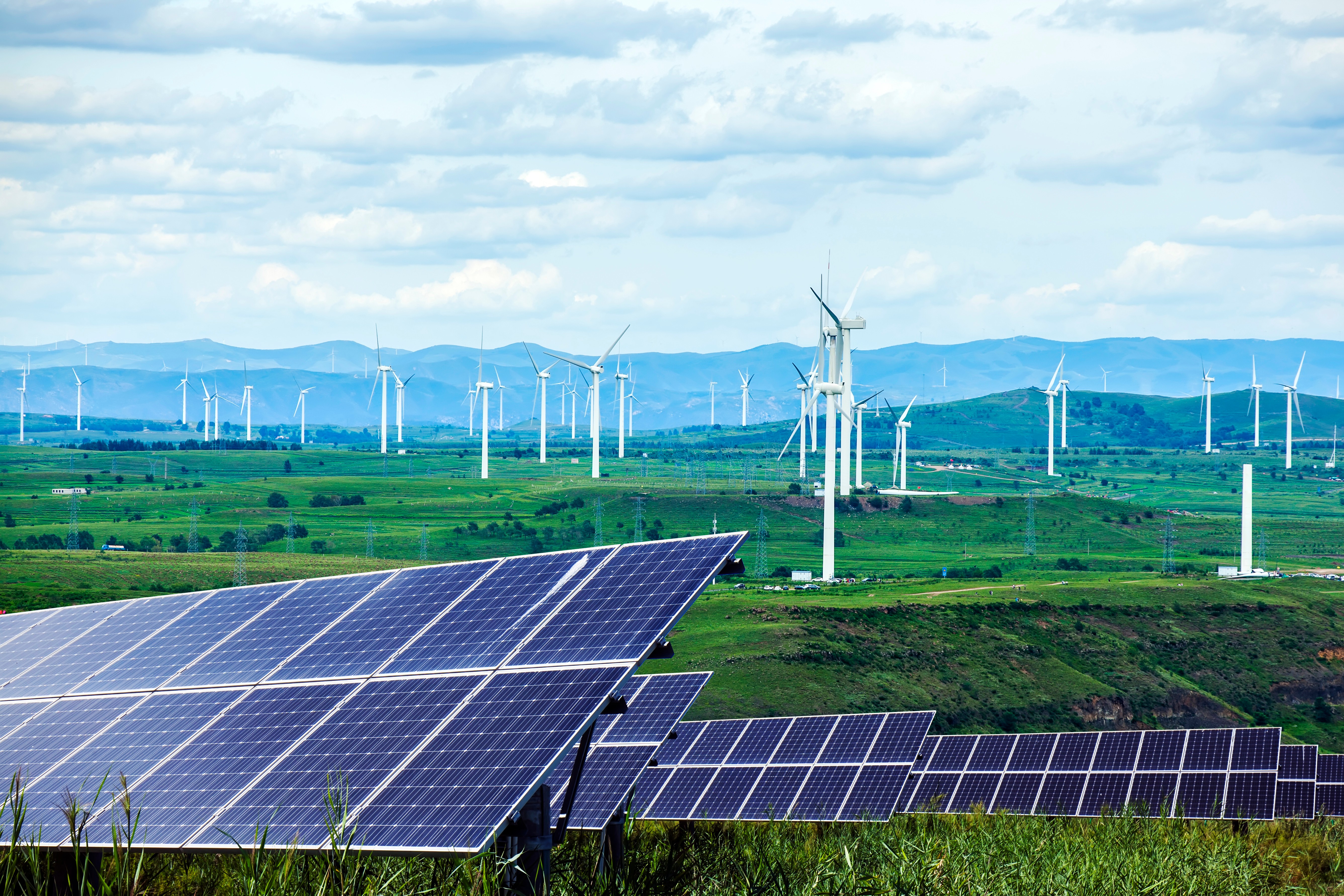 Solar panels and wind turbines in a green landscape with mountains in the background, representing renewable energy sources