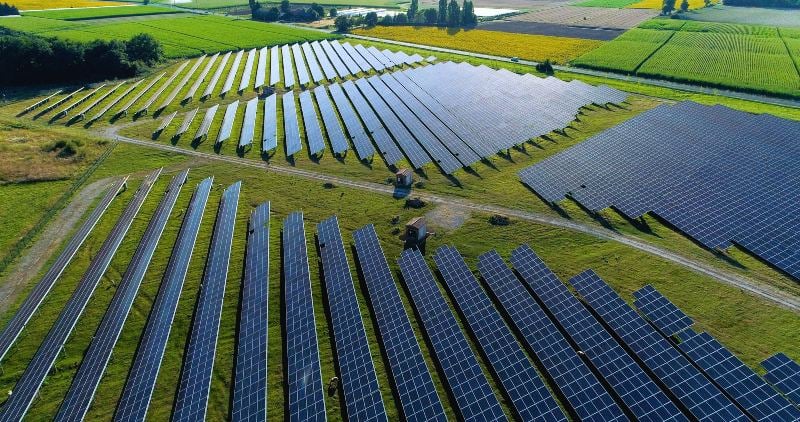 Aerial view of solar panels arranged in rows on green fields, surrounded by agricultural land.