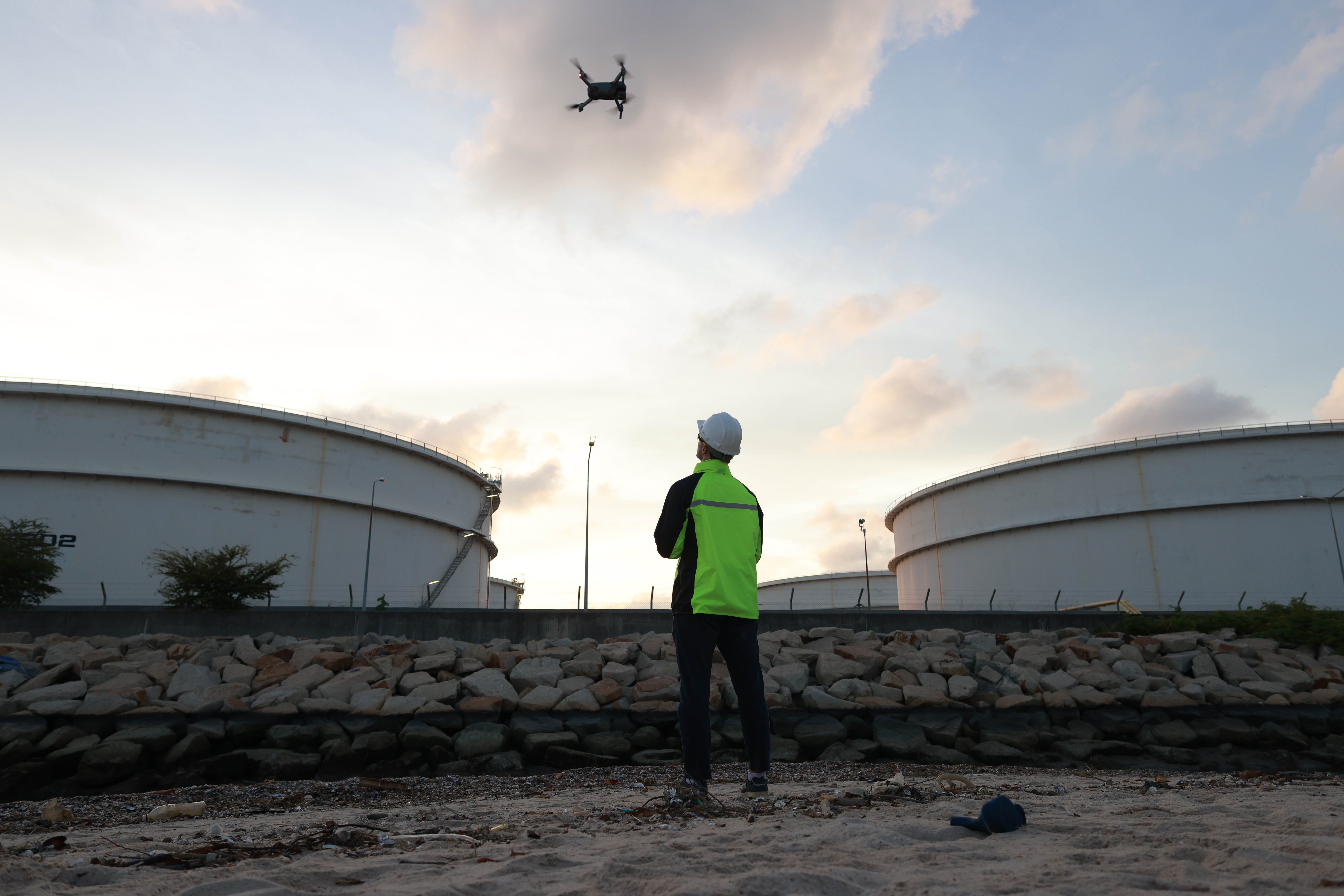 A Kiwa drone pilot during a drone inspection, in a high-visibility jacket and helmet operating a drone near large industrial storage tanks at sunset