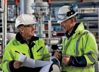 Två män klädda i varseljackor och skyddshjälmar samtalar med varandra i industrilokal.