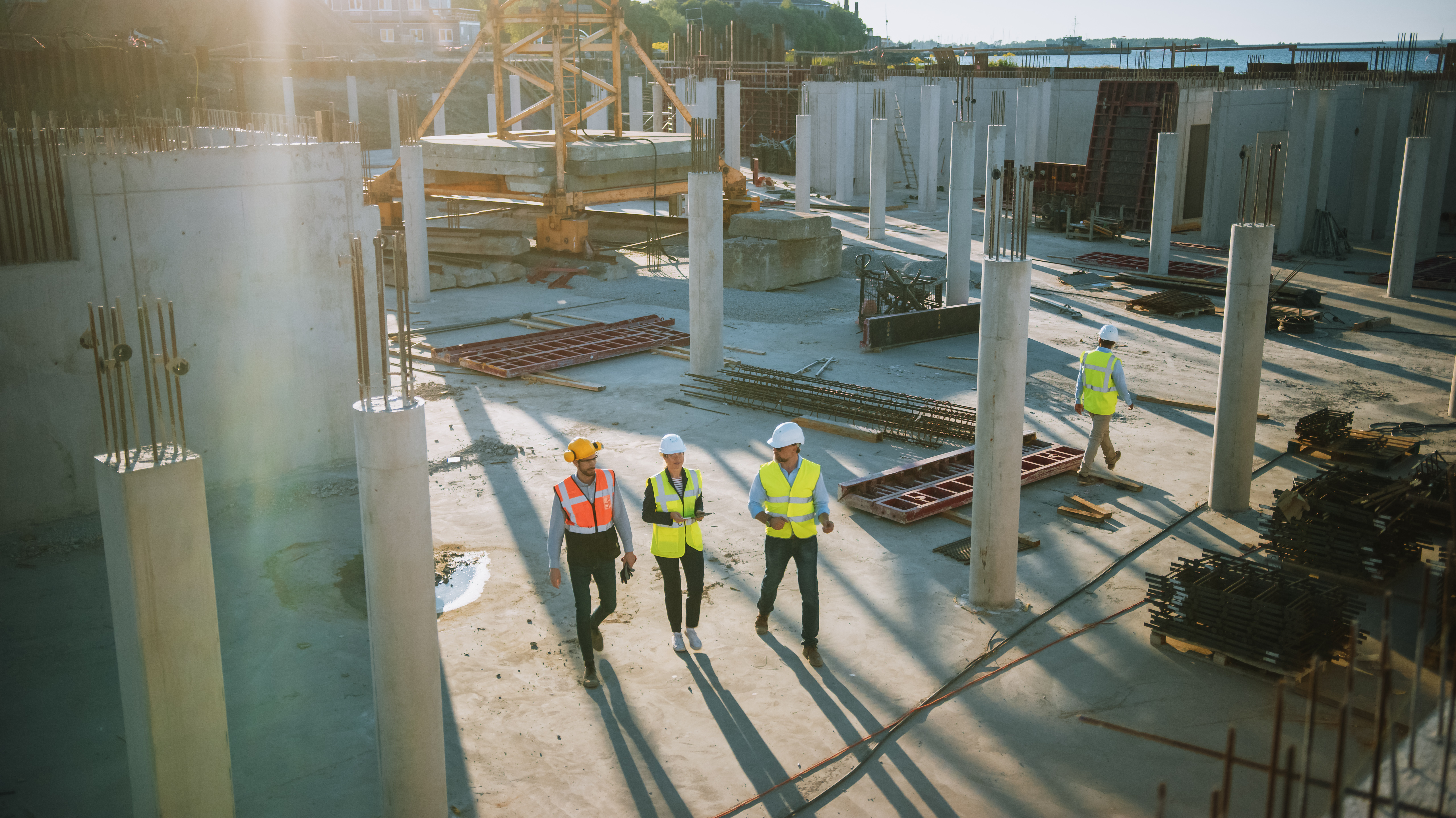 Construction site with four Kiwa employees wearing safety gear and helmets, walking among concrete pillars and construction materials under sunlight