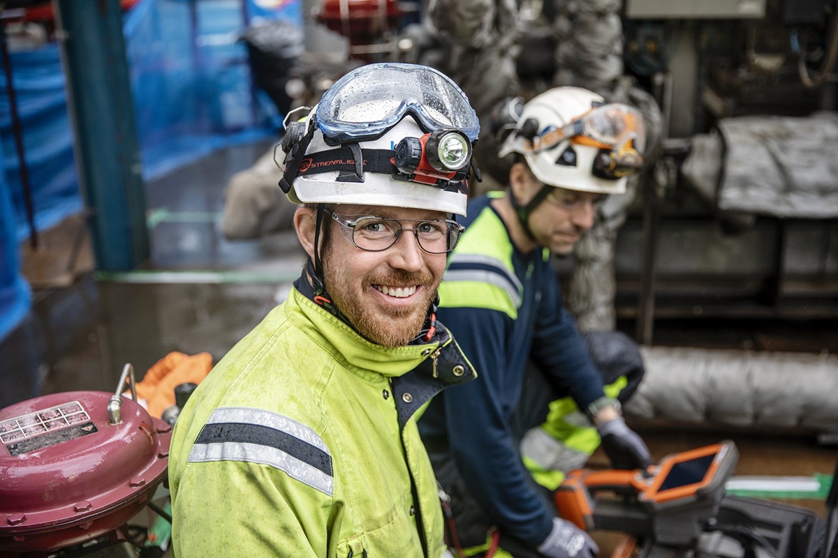 Two workers wearing high-visibility jackets and protective helmets with lights, operating equipment in an industrial setting.
