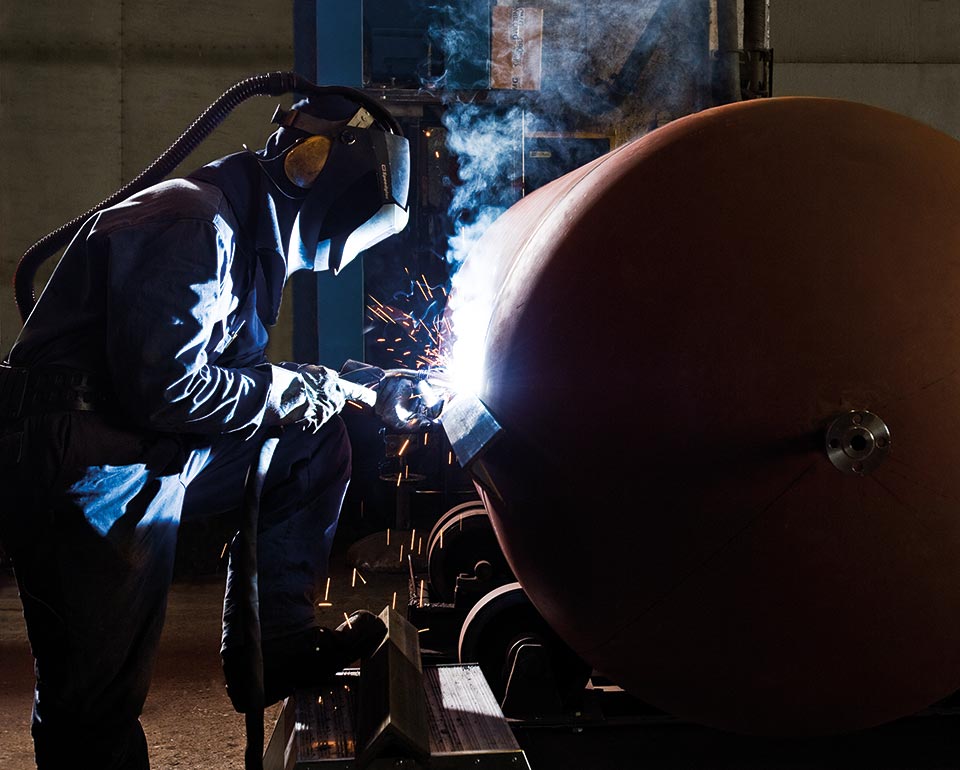 A Kiwa certified welder wearing protective gear performing welding on large metal tank in industrial setting, with visible sparks and smoke