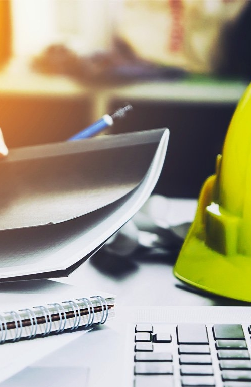 En gul bygghjälm ligger på ett skrivbord bredvid en dator och pappersdokument.