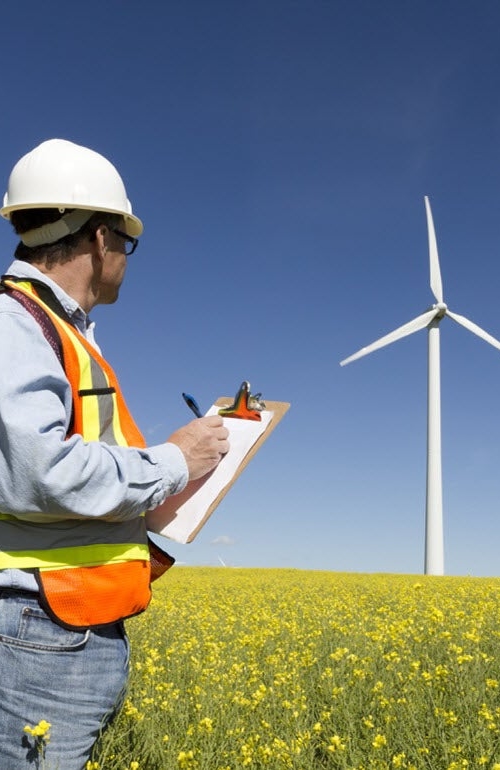 En man i varselväst och hjälm håller i dokument och skriver anteckningar framför en rad med vindkraftverk.