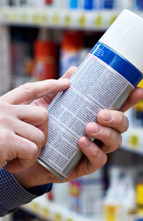 Närbild på händer när en person håller i en sprayflaska och för ett finger längst den finstilta texten på flaskans baksida för att lättare kunna läsa vad som står.