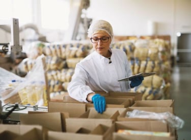 En kvinna, med hårnät, plasthandskar och vit rock, tittar ner i en förpackning med livsmedel.