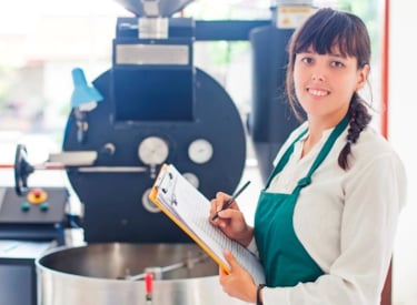 En leende kvinna i förkläde står med en penna och papper bredvid en maskin för livsmedel.