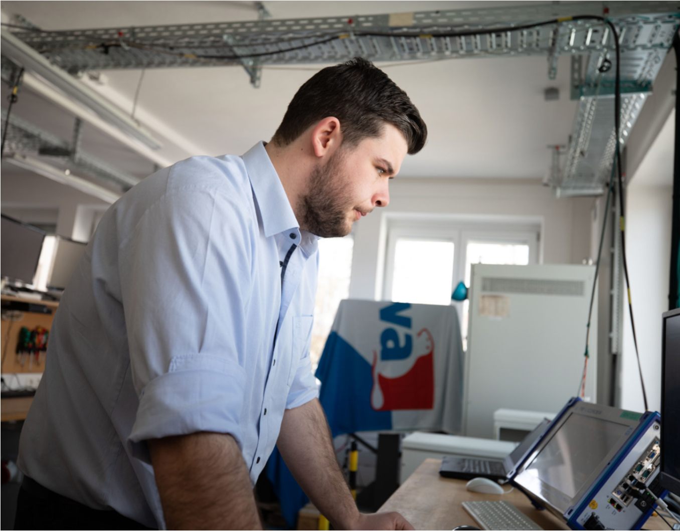 Kiwa lab employee in a laboratory setting focusing intently on a computer screen, wearing a light blue shirt; Lab filled with technical equipment