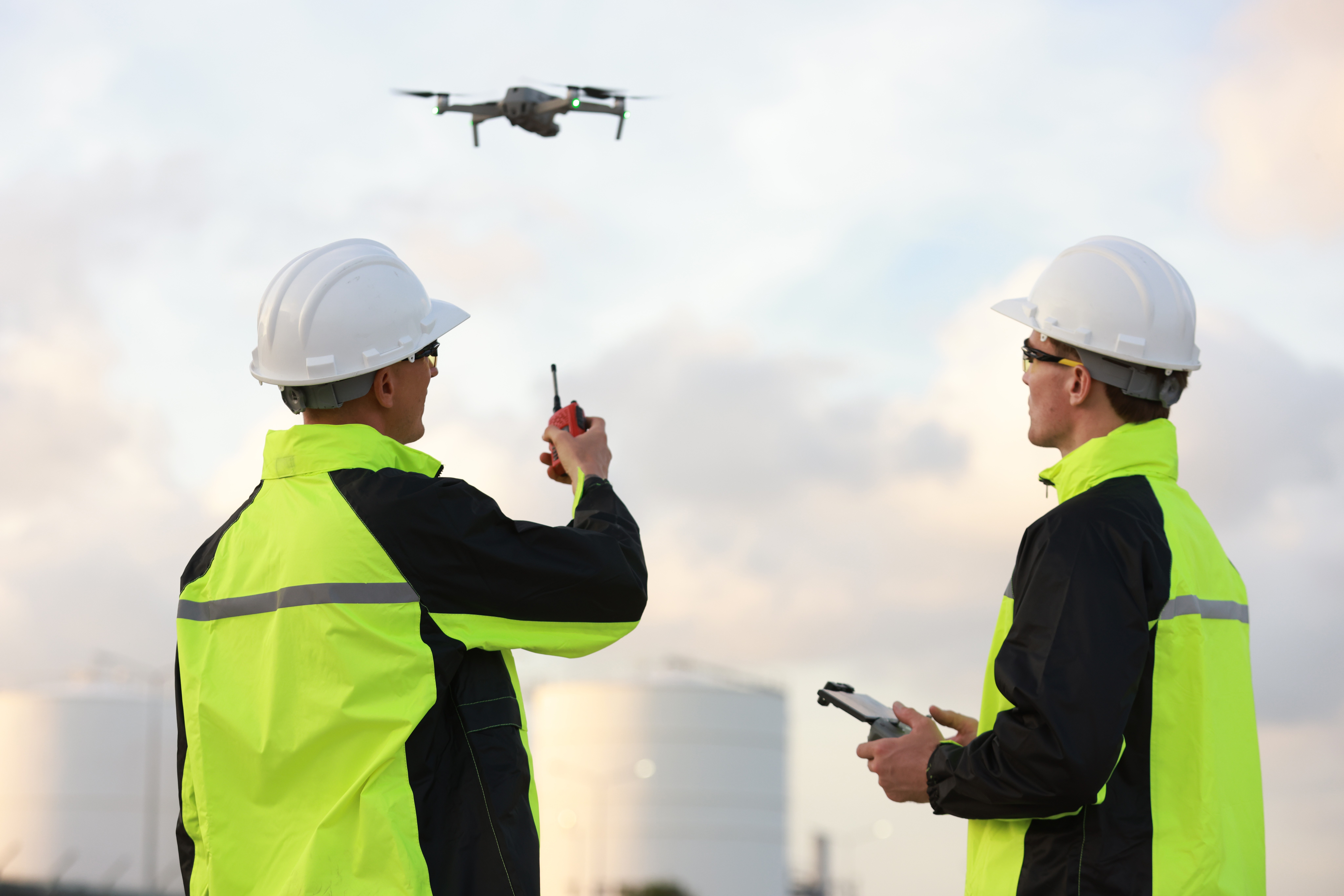 Two workers in white hard hats and high-visibility jackets operate a drone with remote controllers at a worksite.