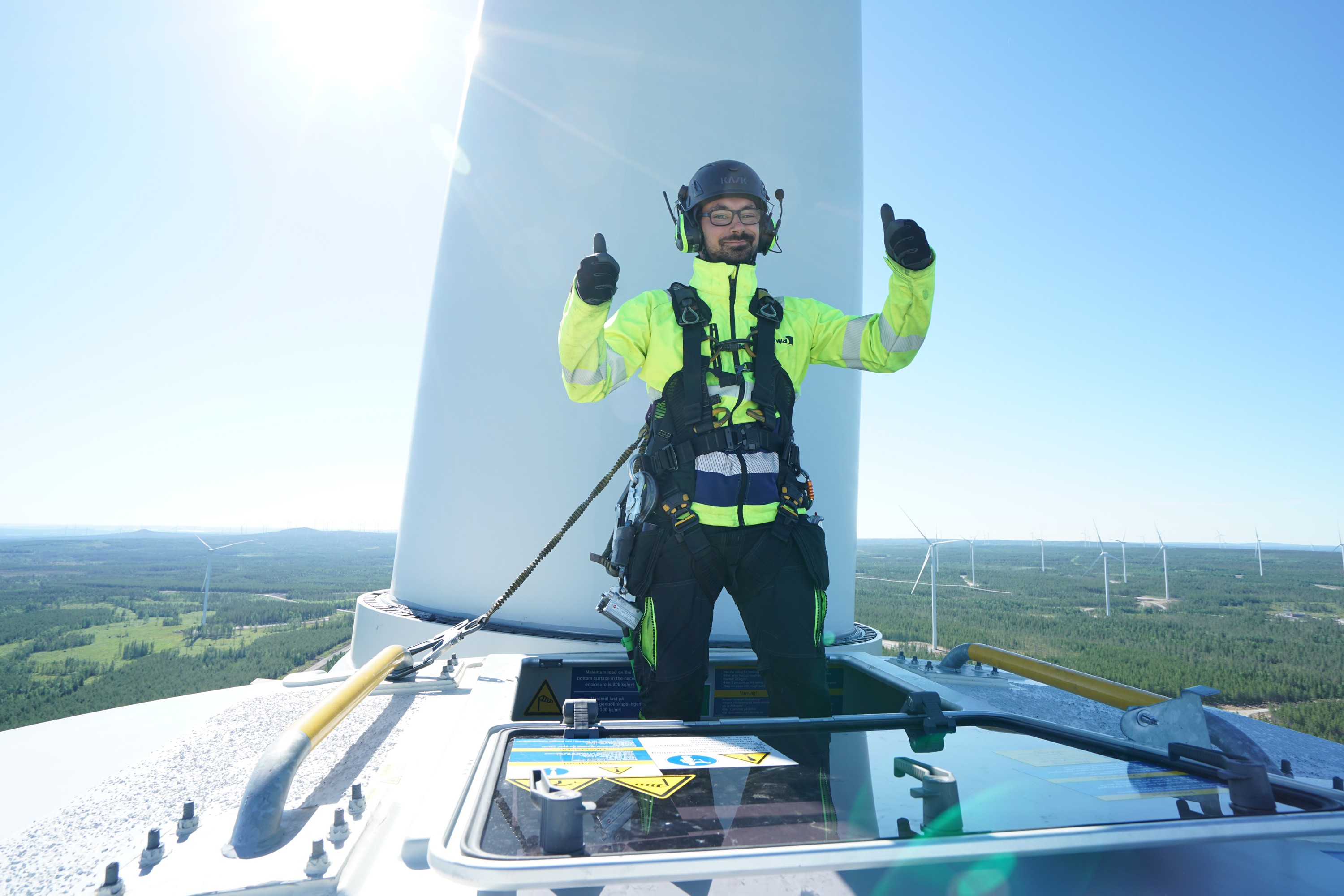 A wind turbine technician in safety gear stands atop a turbine tower giving two thumbs up with wind turbines and a rural landscape in the background.