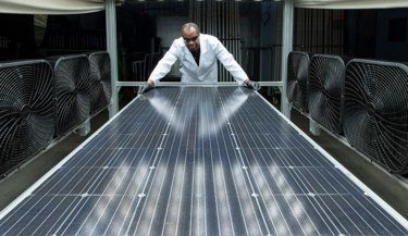 Kiwa laboratory employee in a lab coat inspecting a large solar panel surrounded by industrial fans in a testing facility