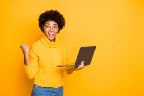 Woman in a yellow sweater excitedly holding a laptop against a bright yellow background, expressing success and positivity