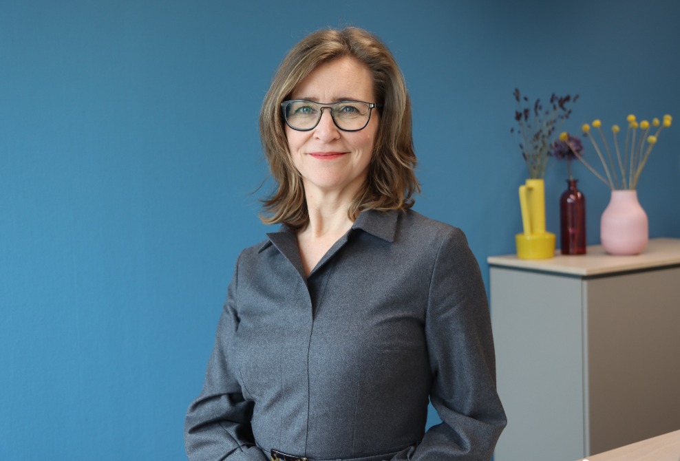 Ivonne, former Kiwa employee, in gray dress with glasses standing in modern office with blue wall and colorful vases