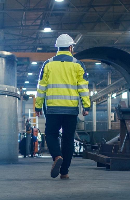 En entreprenadbesiktningsman promenerar bort, klädd i varseljacka och bygghjälm, och ser sig omkring på en konstruktionsplats.