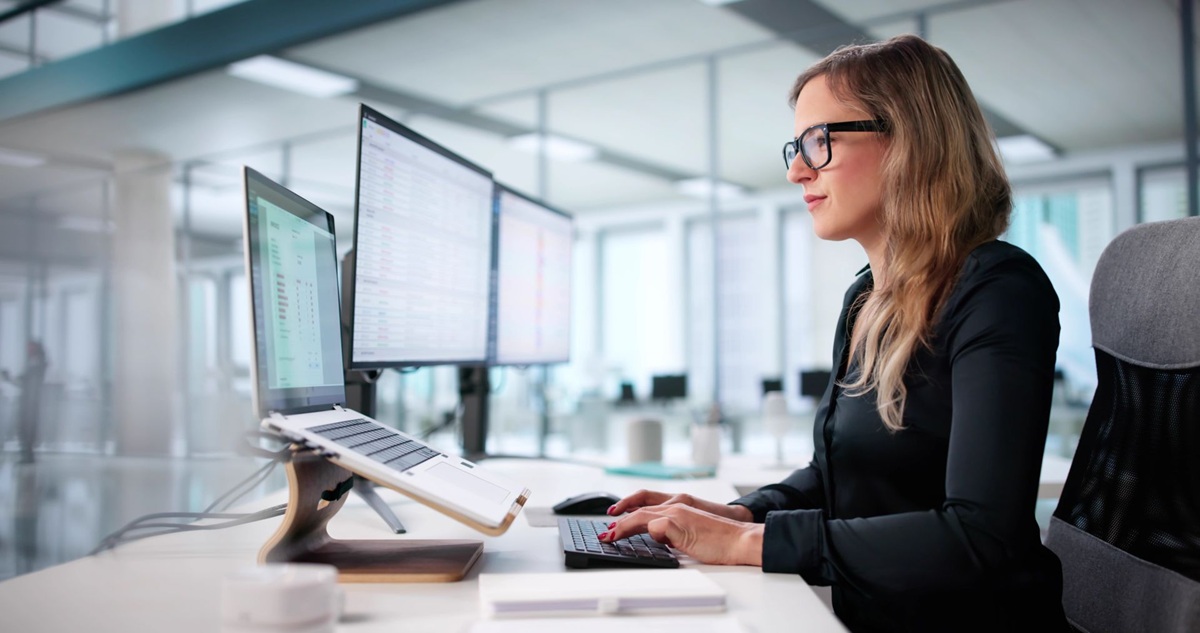 Vrouw in een kantooromgeving typt op een toetsenbord achter meerdere computerschermen aan een bureau.