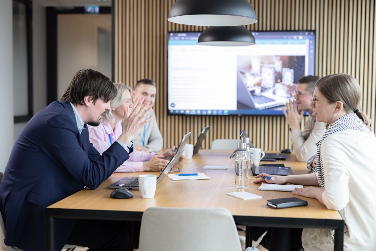 A group of people sitting at a conference table in a modern office, discussing work with a presentation displayed on a screen in the background.