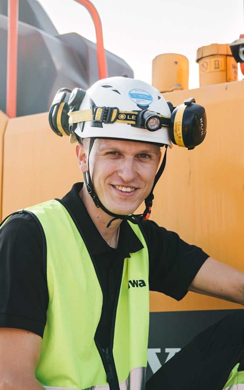 Kiwa inspektör med hjälm och gul varselväst står framför gula grävmaskiner.