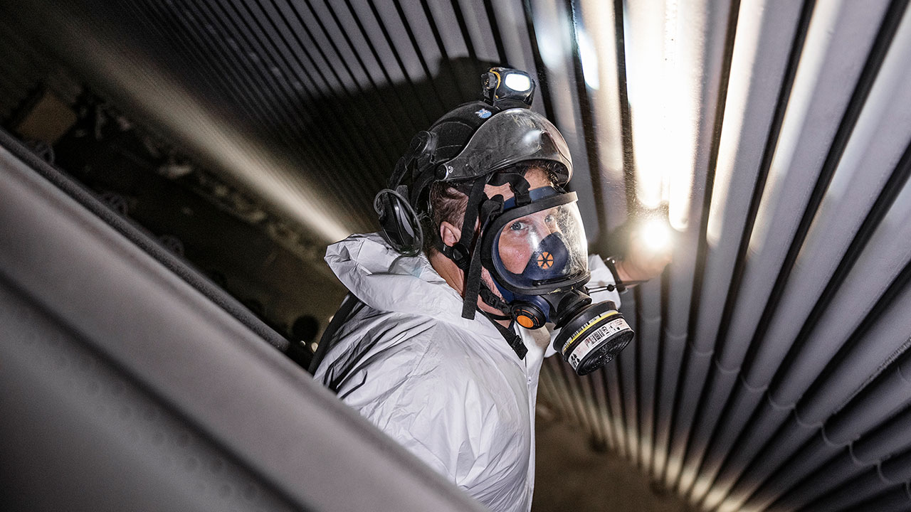 Besiktningsingenjör med gasmask, hjälm och skyddskläder kontrollerar rörledningar för att säkerställa att säkerhetsstandarder efterlevs. 