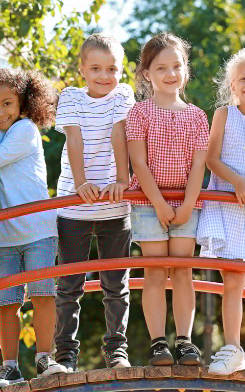 Glada somriga barn står uppställda på rad på en liten gångbro i en lekpark.