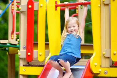 En liten flicka har roligt på en lekplats, hon håller båda händerna i en stång ovanför hennes huvud, hänger lite i den, och står barfota på rutschkanan som hon verkar vilja åka i.