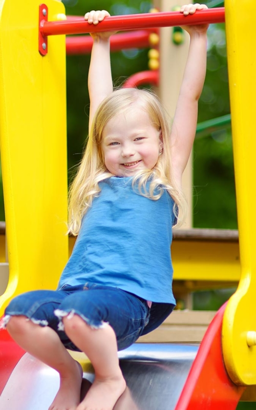 En liten flicka har roligt på en lekplats, hon håller båda händerna i en stång ovanför hennes huvud, hänger lite i den, och står barfota på rutschkanan som hon verkar vilja åka i.