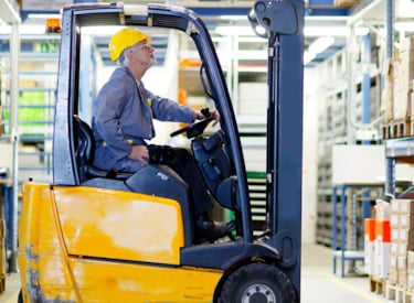 Truckförare kör en truck i ett lager, tittar upp mot pallställ för att lyfta upp varor.