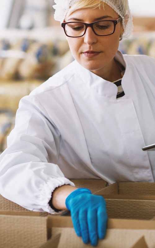 En kvinna med hårnät, skyddskläder och -handskar kontrollerar kartonger med livsmedel.