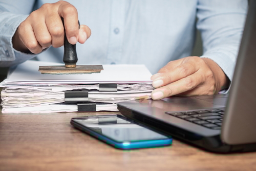 Pappershög bredvid dator på skrivbordet, som är på väg att få en stämpel.
