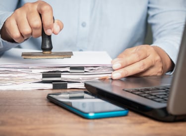 Pappershög bredvid dator på skrivbordet, som är på väg att få en stämpel.