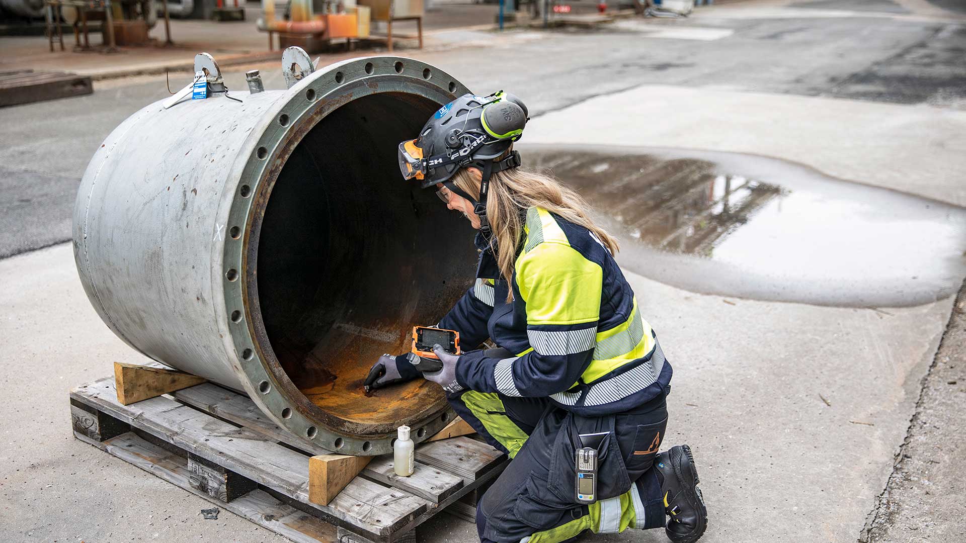 Arbetare i skyddsutrustning inspekterar insidan av ett stort metallrör på en industriell arbetsplats.