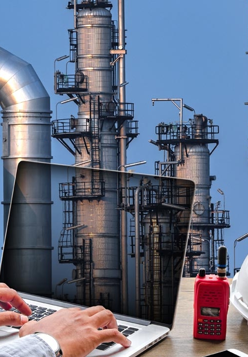 Ingenjör arbetar med dator med skyddshjälm bredvid, gas- och oljeraffinaderi visas i bakgrunden.