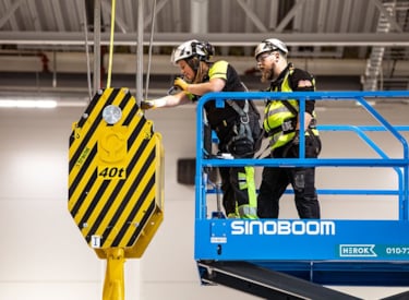 Två besiktningsingenjörer befinner sig högt upp i en mobil arbetsplattform där de kontrollerar en krananordning.