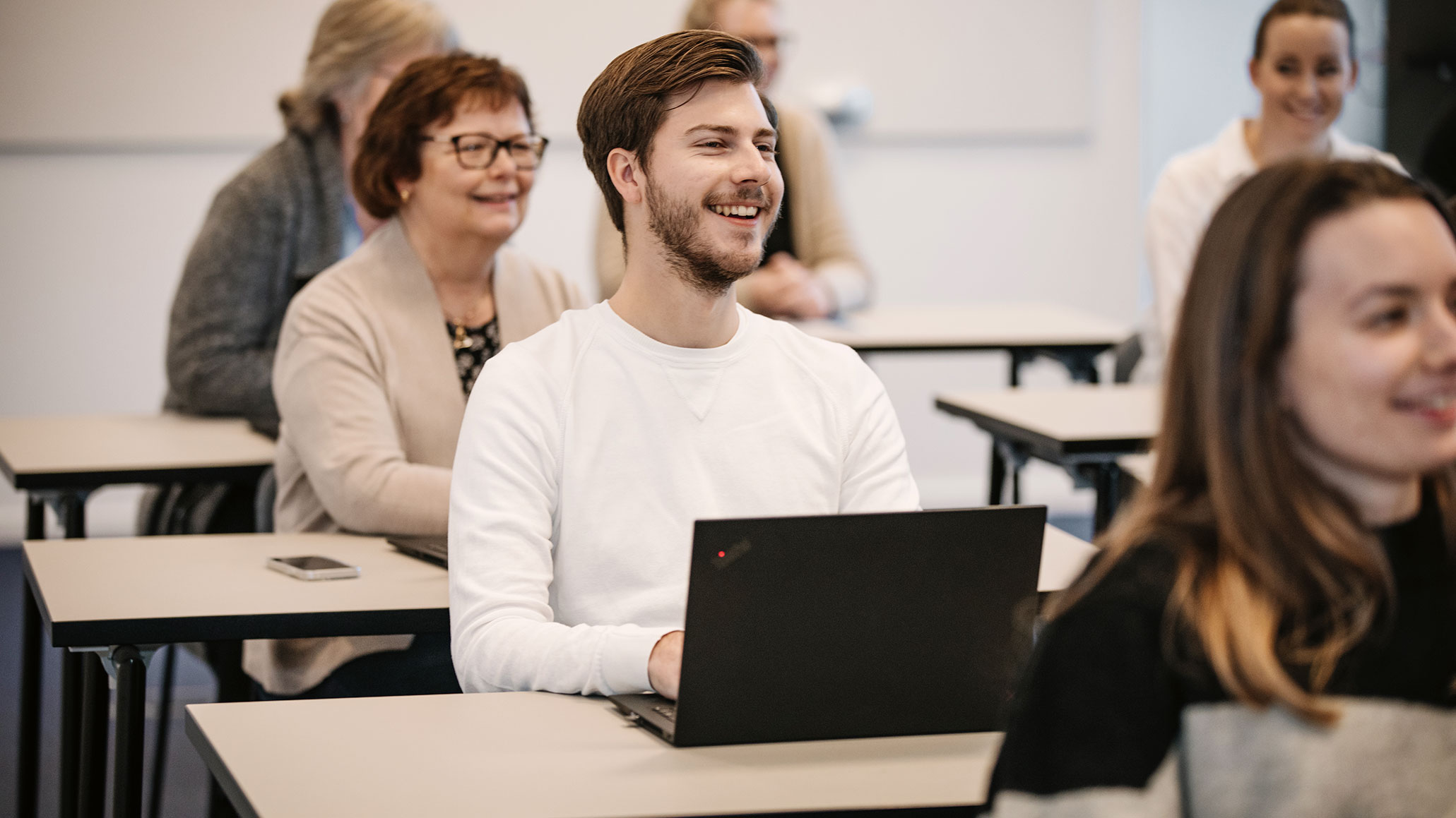 Vuxna elever i kurslokal som sitter vid bord med en dator framför sig. 