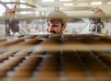 En leende man rullar en ställning med flera bakplåtar fulla med bröd på ett bageri.
