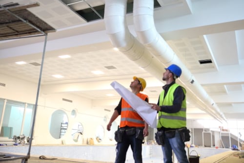 Kiwa auditors in safety gear inspecting ceiling ducts in an indoor facility, reviewing blueprints for building maintenance and HVAC system installation