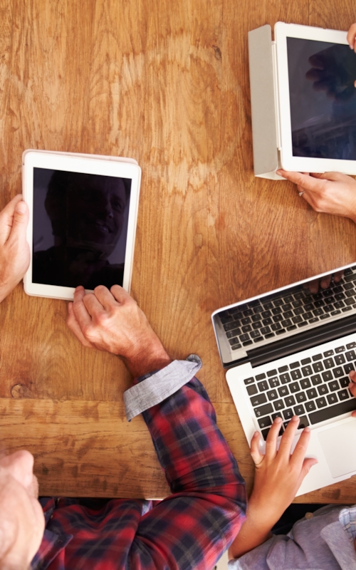 Family of 4 sitting around a table seen from above. All of them using mobile devices like phones, tablets and laptops that have been EMC tested by Kiwa