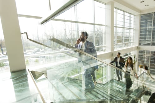 Modern office interior with business professionals walking up a glass staircase, featuring large windows and natural lighting