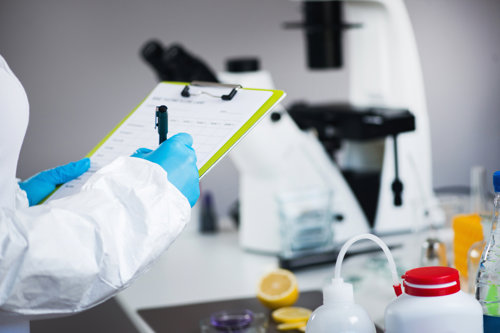Scientist in lab coat and gloves recording data on clipboard in laboratory with microscope and equipment