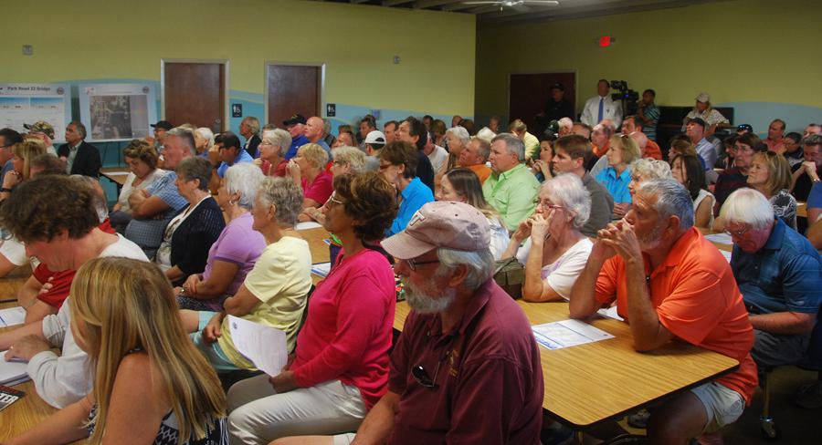 Community members gather in a meeting room for a local public discussion, seated at tables and attentively listening to speakers