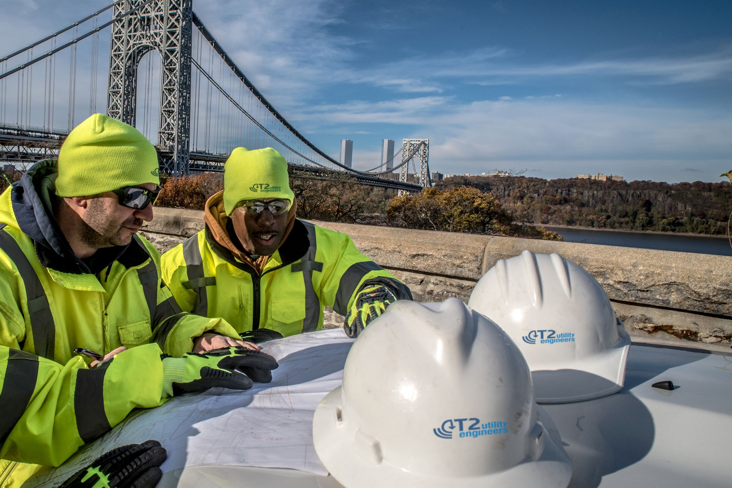 Kiwa T2UE engineers in high-visibility jackets and helmets reviewing plans near a bridge on a clear day