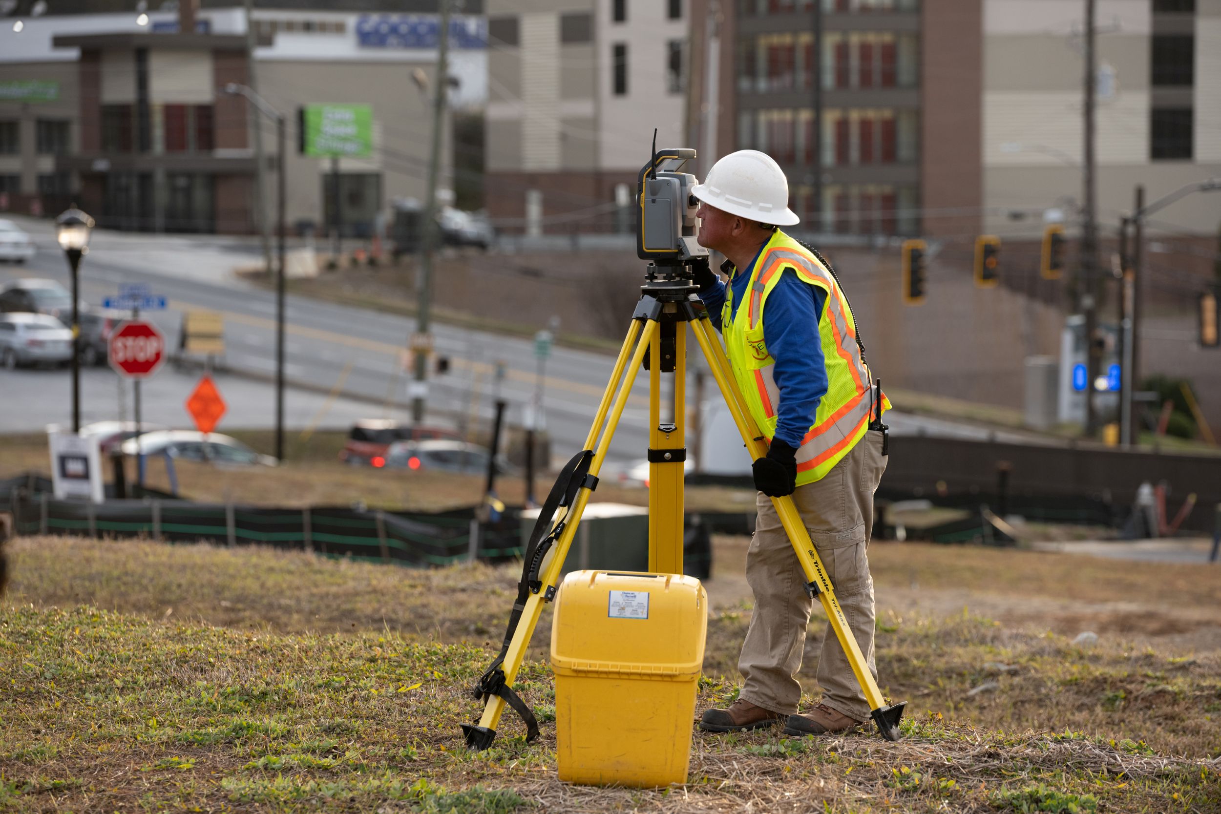 Construction surveyor using a tripod-mounted total station on a roadside site, wearing a hard hat and reflective safety vest.