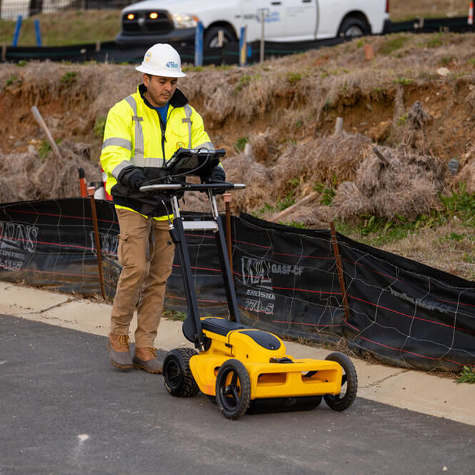 Kiwa T2UE employee in a high-visibility jacket and hard hat using a ground-penetrating radar cart at a roadside work site.