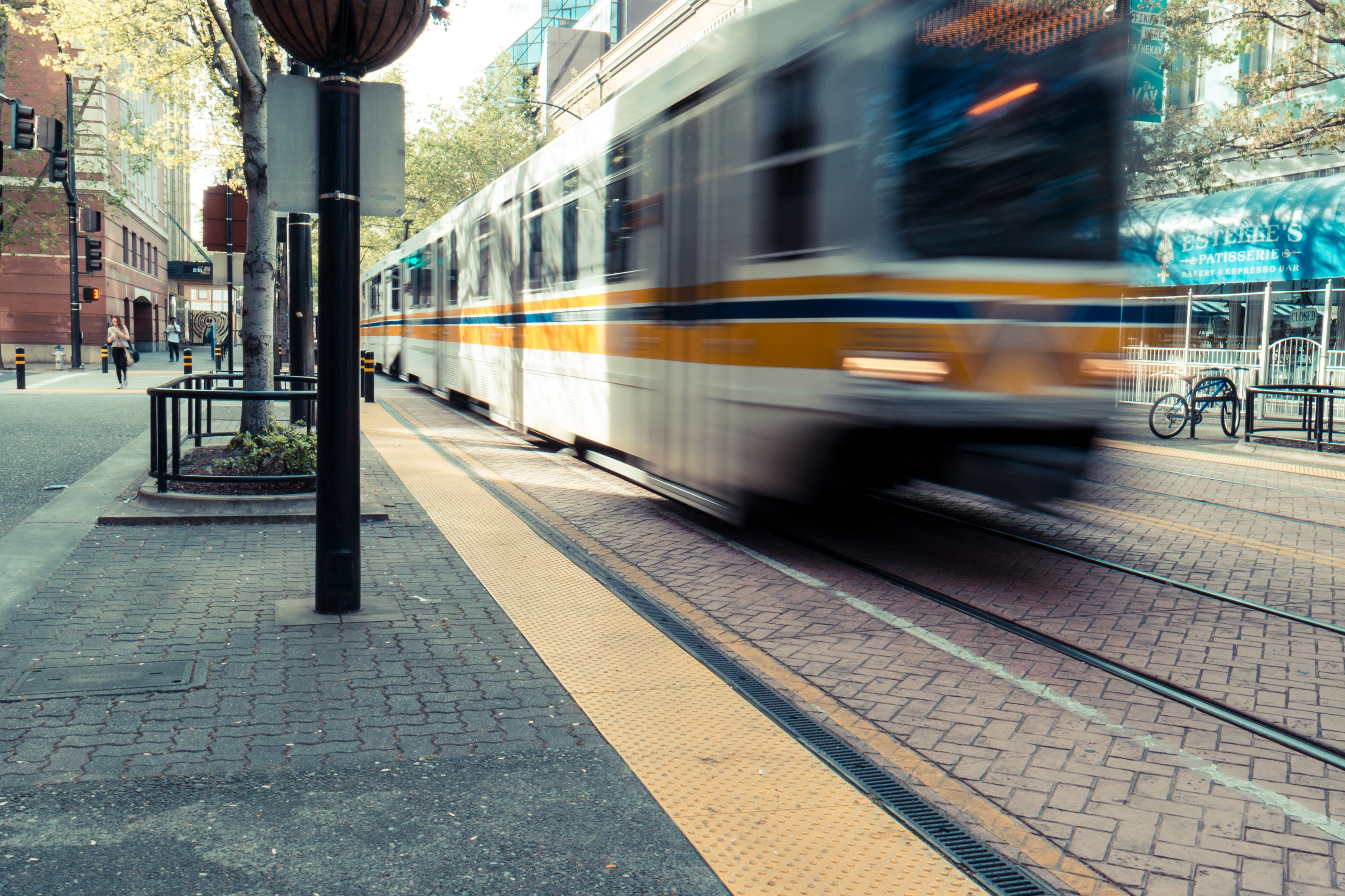 A blurred image of a moving tram on a city street, with buildings, trees, and a bicycle rack in the background.